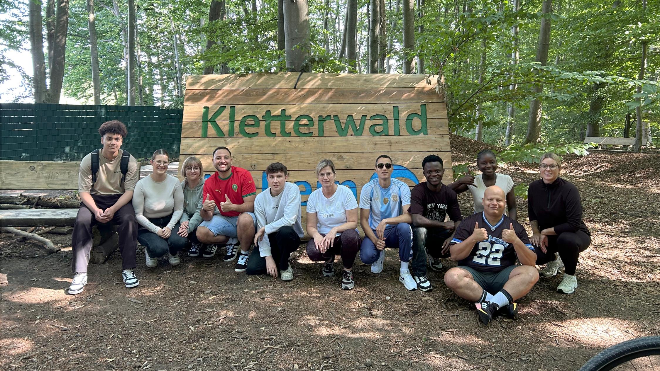 11 Auszubildende zur Pflegefachfrau beziehungsweise zur Pflegefachfrau knien vor einem Schild, auf dem Kletterwald steht.