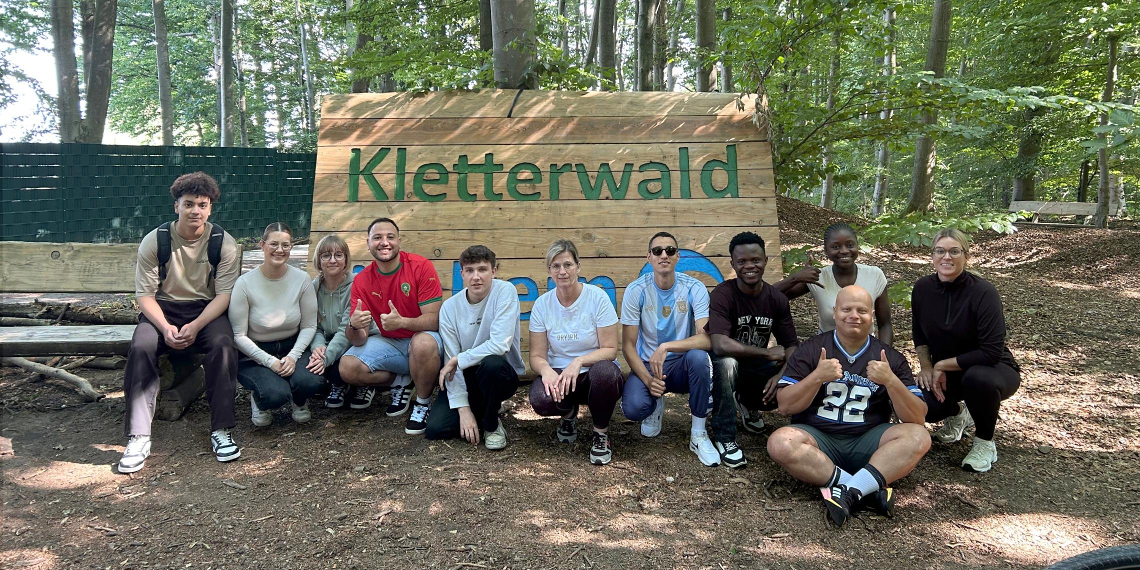 11 Auszubildende zur Pflegefachfrau beziehungsweise zur Pflegefachfrau knien vor einem Schild, auf dem Kletterwald steht.