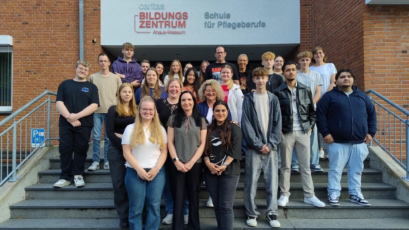 27 angehende Pflegefachassistentinnen und Pflegefachassistenten stehen auf mehreren Treppenstufen der Schule für Pflegeberufe und schauen in die Kamera.