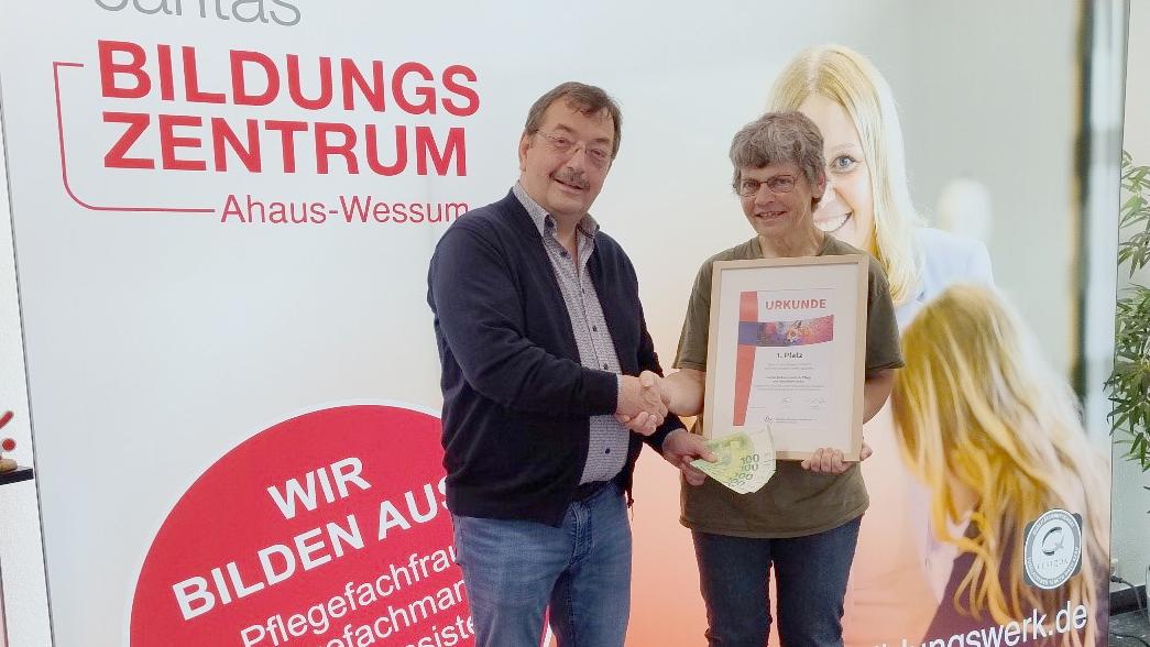 Schulleiter Reinhard Sicking überreicht vor einem Plakat der Pflegeschule in Ahaus-Wessum Maria Südholt das Preisgeld in Höhe von 500 Euro.