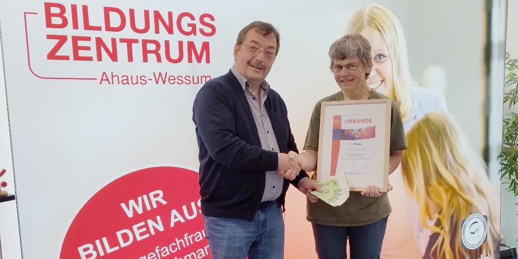 Schulleiter Reinhard Sicking überreicht vor einem Plakat der Pflegeschule in Ahaus-Wessum Maria Südholt das Preisgeld in Höhe von 500 Euro.